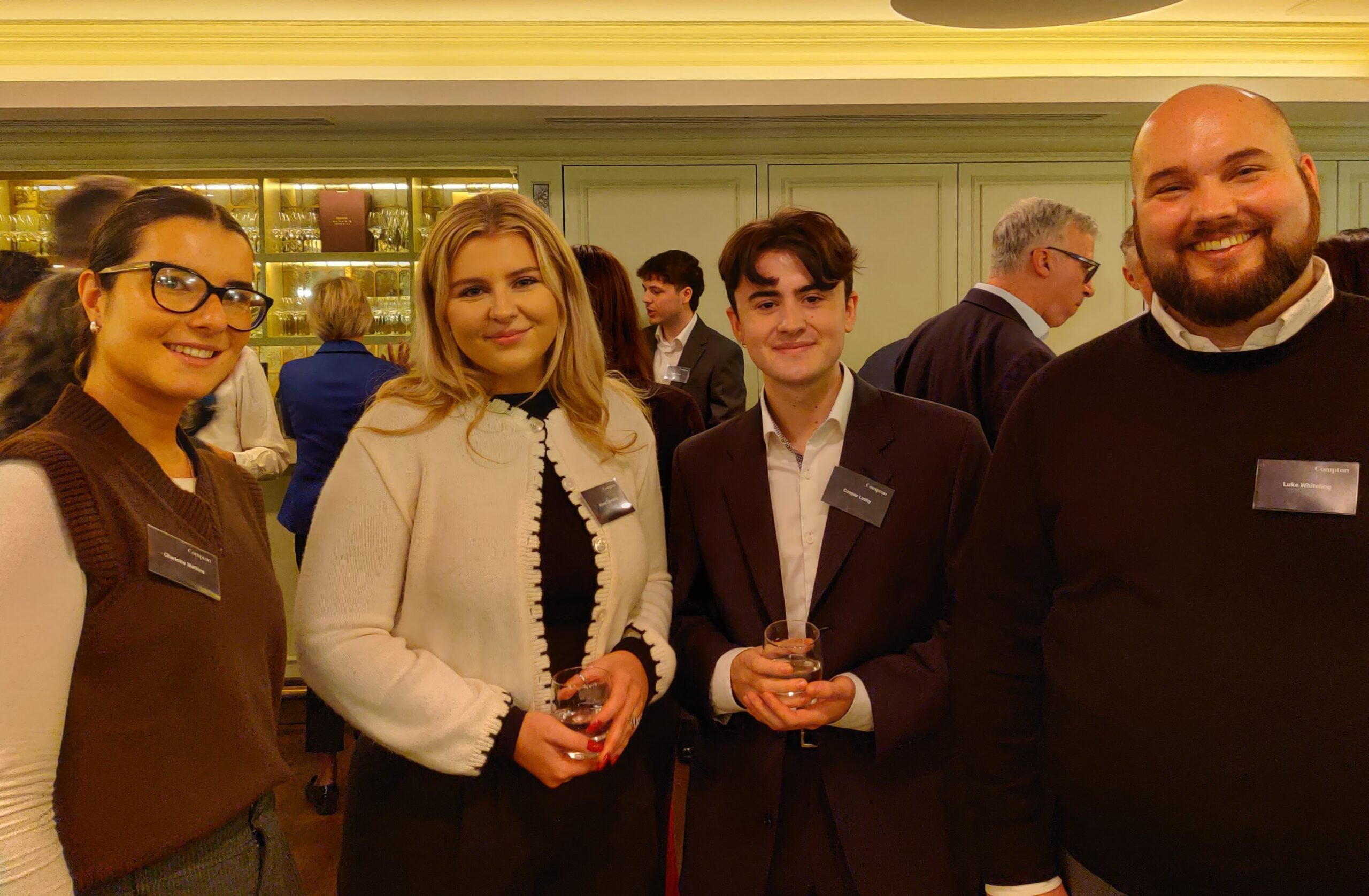 A group of four people standing together at a networking event, each wearing name badges and holding drinks. They are indoors, in front of a wall with glass shelves displaying bottles and glassware. Other attendees are visible in the background, engaged in conversation. The lighting is warm and ambient, creating a formal yet relaxed atmosphere.