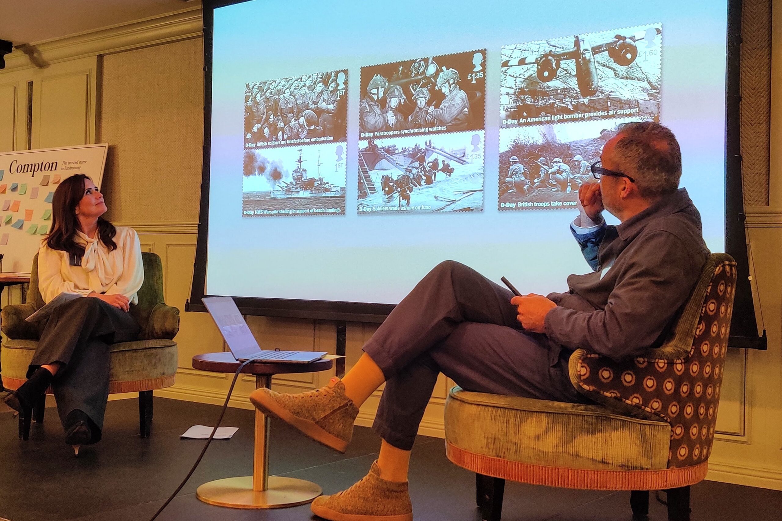 Two people are seated on stage in a discussion setting, with a large screen behind them displaying a series of black-and-white postage stamps featuring historical wartime images. One person is sitting on the left in an armchair, while the other is on the right, leaning back in a patterned chair and holding a pen. A small round table with a laptop is positioned between them. A flip chart with the word “Compton” is visible in the background.