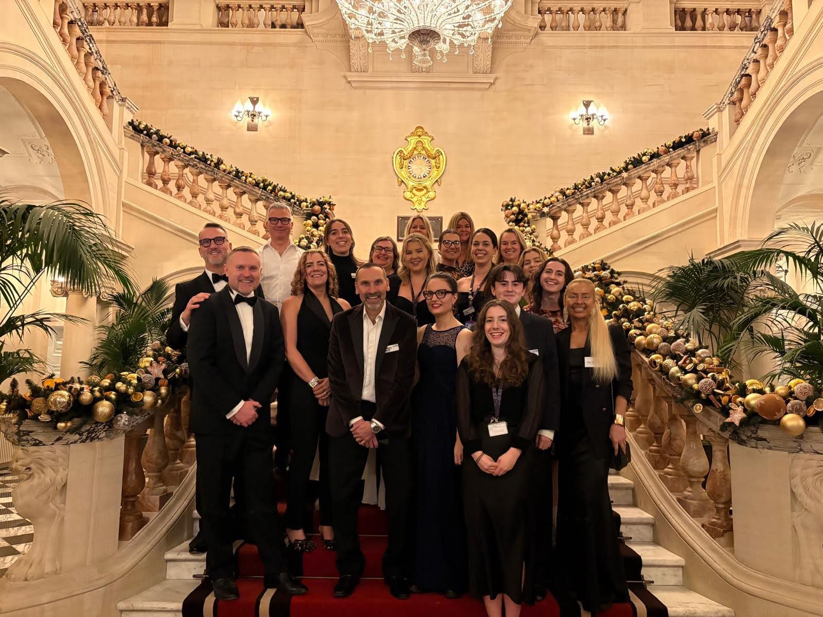 A large group of people dressed in formal evening attire standing on a grand staircase inside an opulent venue. The staircase is adorned with festive garlands featuring gold and black ornaments, and a red carpet runs down the center. The background showcases an ornate chandelier, decorative wall sconces, and a golden crest mounted on the wall, adding to the luxurious setting.