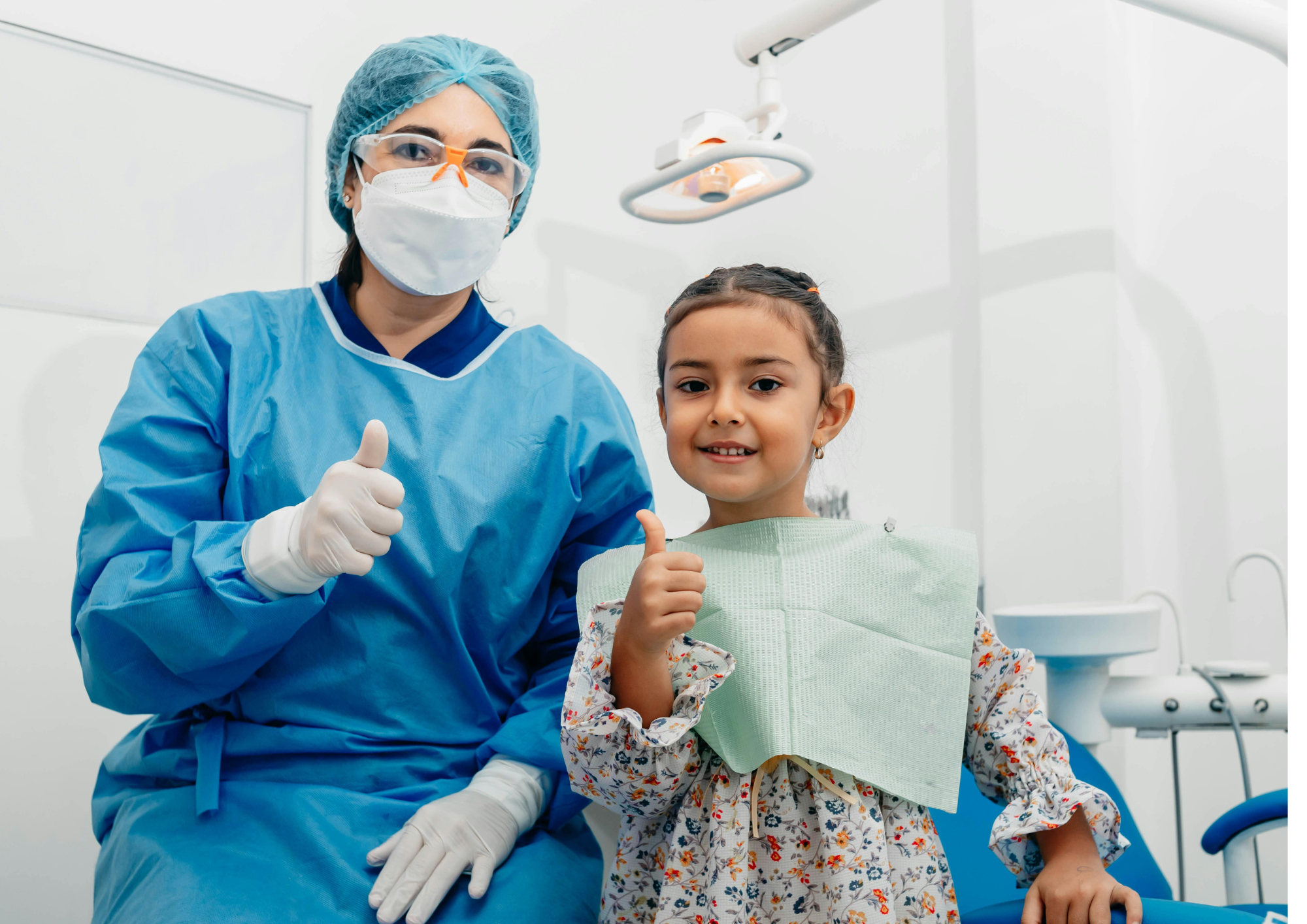 A dental professional wearing protective clothing, gloves, face mask and safety glasses sits beside a child patient in a dental clinic. Both are giving a thumbs-up gesture. Dental equipment and an examination light are visible in the bright clinical setting.